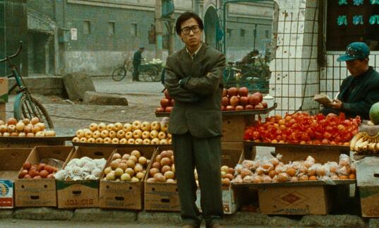 A man stands in front of a fruit stall with his arms folded across his chest.
