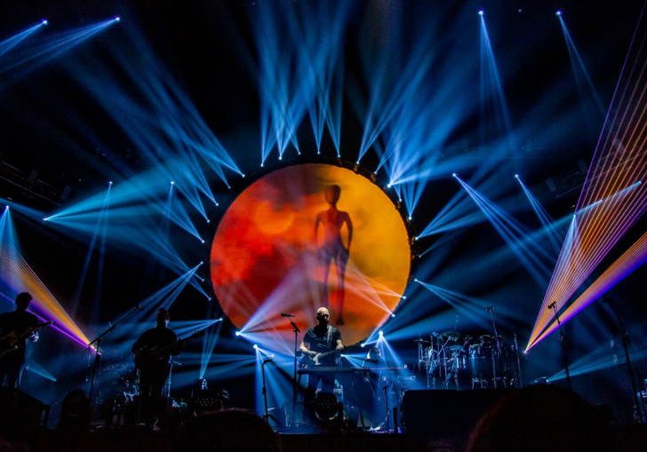Brit Floyd performing on stage, with blue lasers and an orange sun.