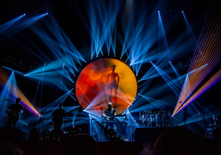 Brit Floyd performing on stage, with blue lasers and an orange sun.