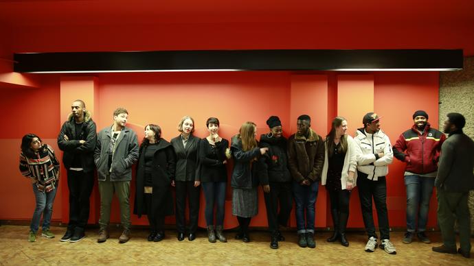 13 people standing in a line along an orange wall, talking and laughing