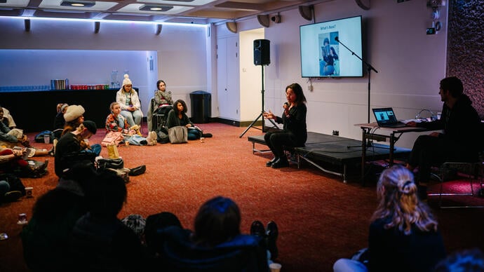 Overview of the room. Group of people sitting on the floor facing a screen with a person standing in front of the screen. 