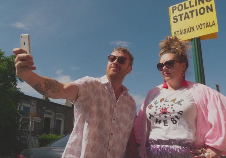 a man and a woman dressed in pink take a selfie with the polling station sign behind them