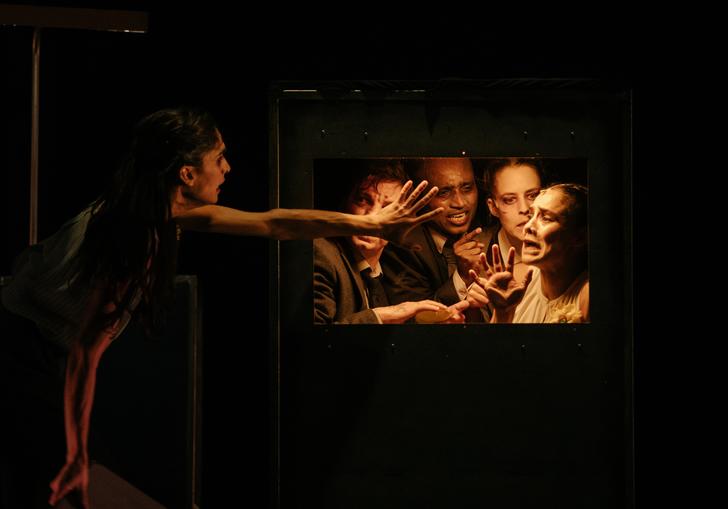 Actors on stage: four people are trapped behind a door, looking desperately at another person reaching towards them.