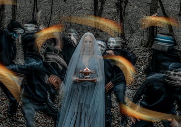 A lady in long kthin light blue veil is staring at the camera. She is holding a small model of a red and white carousel in her hands. Behind her seven masked figure and blurred as they move, they are holding flares. 