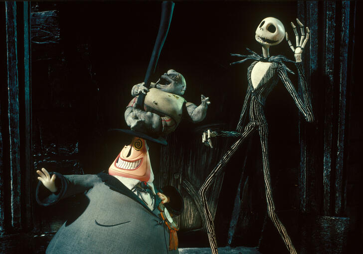 A skeleton man and a short man in a suit dance together on a dark stage.