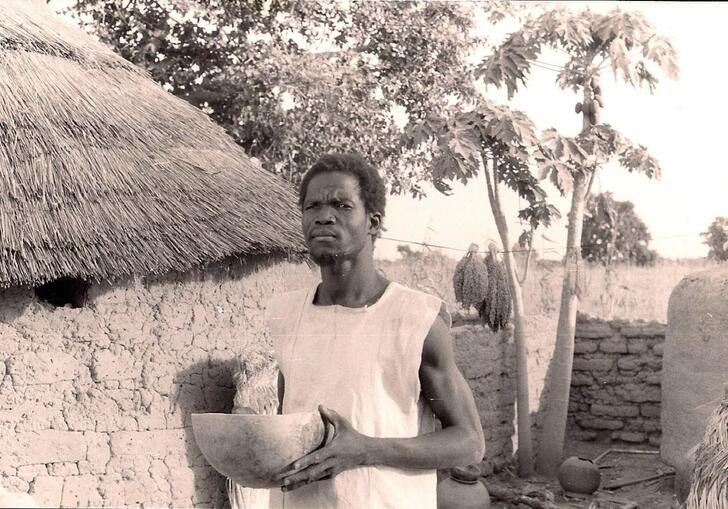 A man stands in front of a thatched roof mud hut, holding a clay pot, with a confused look on his face.
