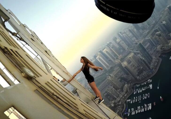 A longhaired woman in business and trainers wear lies back against a skyscraper in front of a city skyline. 