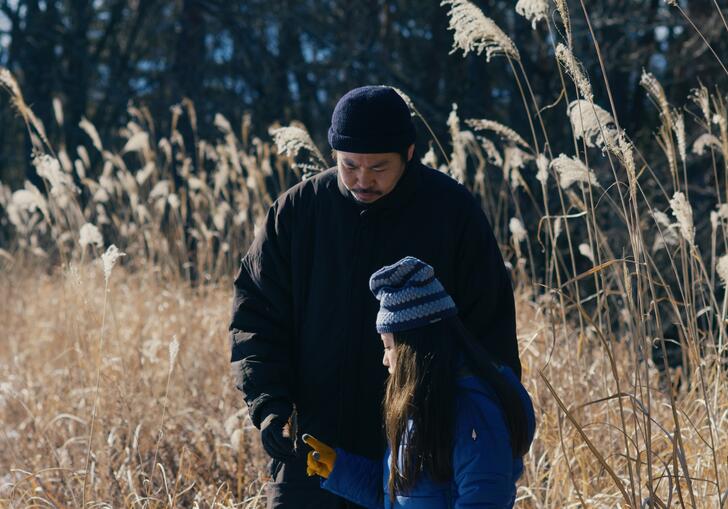 A man stands with his young daughter at the edge of a pond, both dressed in winter clothing. 