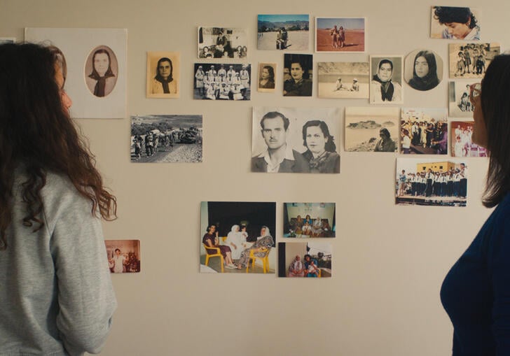 Two women look at a wall that has photographs and notes pinned on to it. 