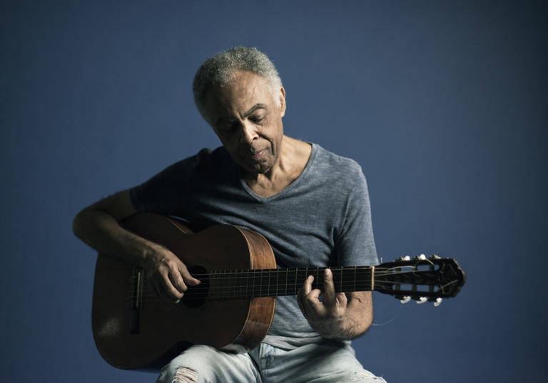 Gilberto Gil looking blue whilst playing his guitar in blue jeans and a blue t-shirt in front of a blue background