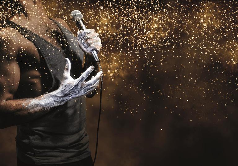 Gold image with a man in black vest top holding a microphone and throwing glitter. 