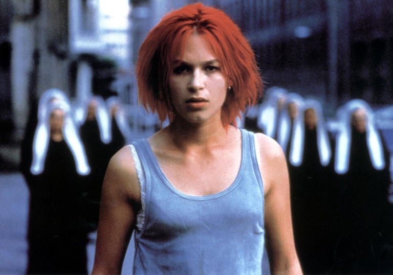 Franka Potente has bright red hair and a grey tank top, standing in front of some blurry nuns in Run Lola Run