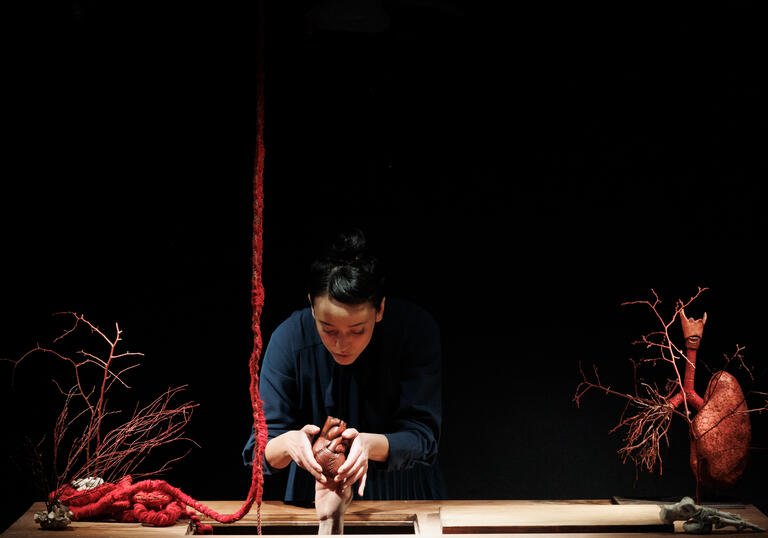 A person stands at a wooden work bench and moulds a small human body out of clay. A large red rope hangs from the ceiling like intestines. 
