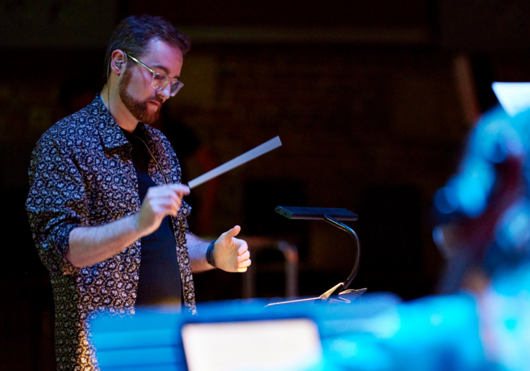 Composer Anselm McDonnell conducing with a baton. He has a colourful patterned shirt, glasses and red hair.