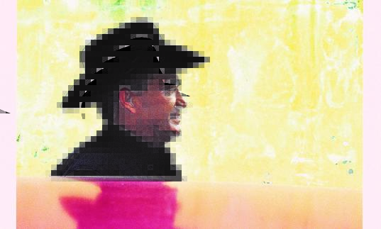 Eliades Ochoa in profile, wearing a black wide-brimmed hat, standing next to a yellow wall