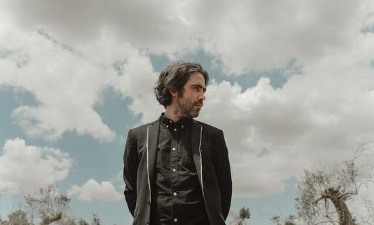 Patrick Watson wearing a black shirt and black jacket looking over his left shoulder, the sky behind him is cloudy