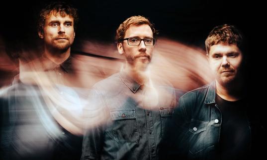 The three members of GoGo Penguin: three white men wearing denim shirts with short haircuts. There is a swirling effect over the image.