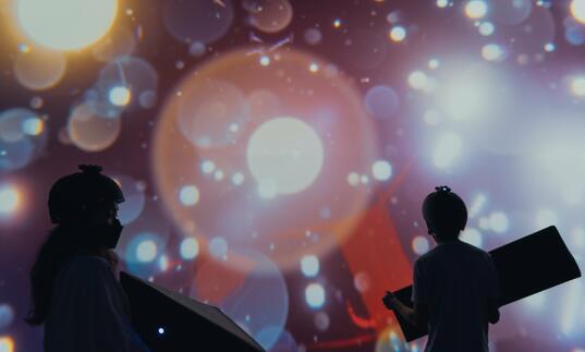 Two silhouettes of a girl and a boy wearing helmets and holding block shapes in front of a digital screen that shows colourful bubbles