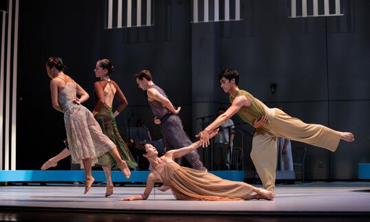 An ensemble of five dancers move and pose elegantly in muted clothing. At the forefront of the stage, two dancers clasp hands. 