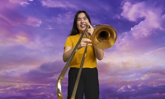 A trombonist from The National Youth Orchestra holding her trombone in front of a skyscape background