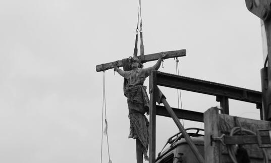 A statue of a saint on a crucifix hangs over a dark sky.