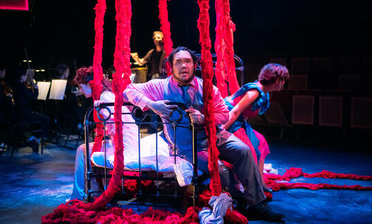 Three opera singers sat on a bed with red rope-like hanging down from ceiling. Small ensemble performing in background