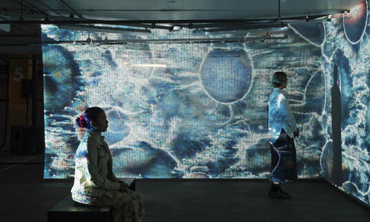 Two people sit and stand, bathed in patterned blue lights being projected onto screens