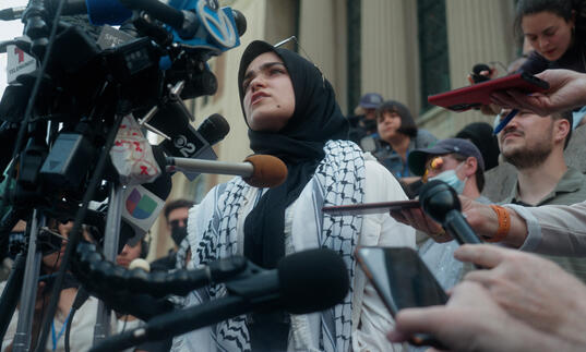 A woman wrapped in a kefiyah speaks into a camera and surrounded by journalists