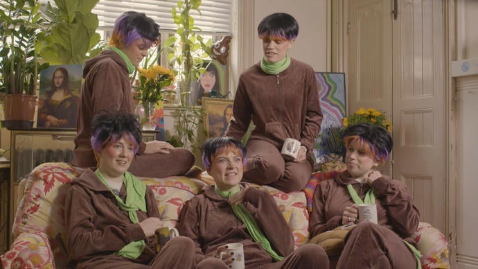 Group of five women sitting on a sofa wearing brown jumpsuits and wigs