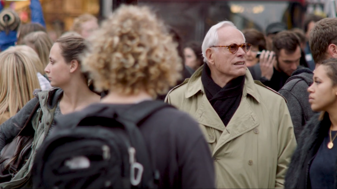 photo of an old man wearing a trench coat standing in a crowd of people