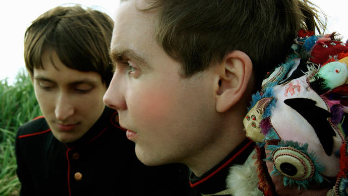 Jonsí & Alex, in military style jackets and feathered adornments, sit in long grass