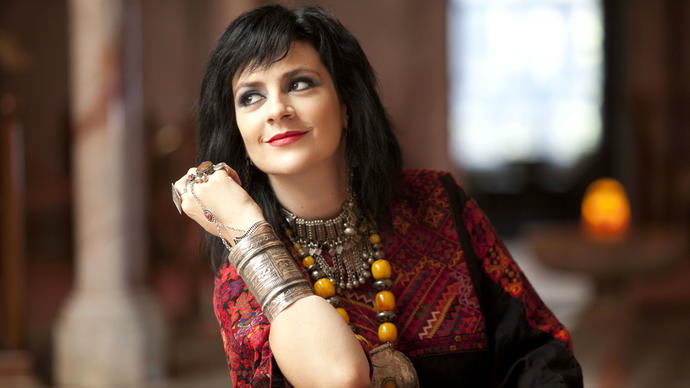 Rim Banna wearing a red dress and large jewellery. She is leaning on her elbow and looking away from the camera.