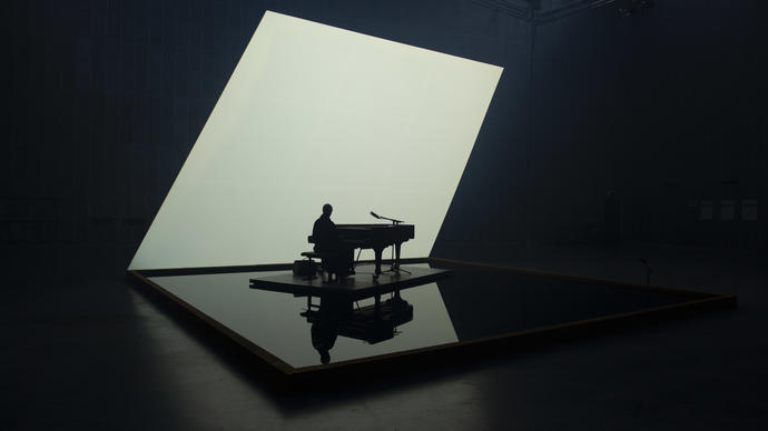 Nik Bartsch playing solo piano on a pool of water, with a lit white backdrop