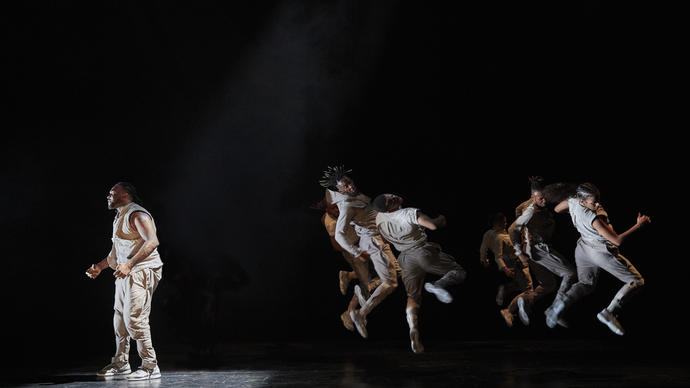 A dancer is looking anguished in the spotlight, while behind them other dancers jump in the air in unison