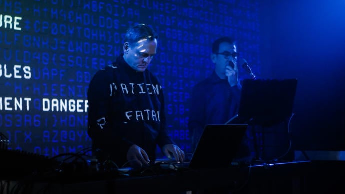 Alva Noto and Anne-James Chaton performing with a laptop and microphone. Words are projected behind them and onto their clothes