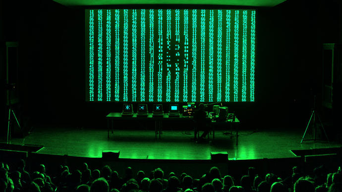 image of live performance shows robert henke standing in front of green and back digital screen with a bank of computers and electronics set up
