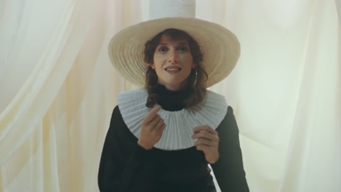 Photo of a lady wearing dark clothing and a big white hat whilst dancing in a white room
