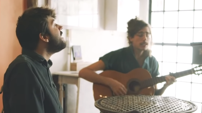 photo of a Tim Bernardes sitting down playing the guitar with Salvador Sobral singing next to him