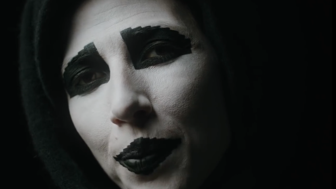 Photo of a lady in a dark space looking very closely into the camera whilst wearing white paint on her face with black filled circle around her eyes.