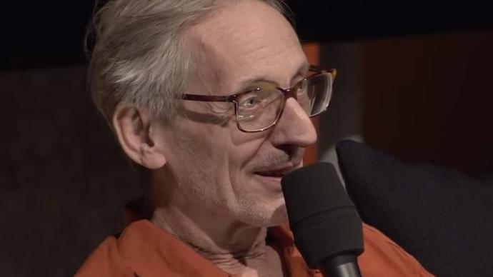 Photo of Manuel Göttsching talking into a microphone, wearing a orange shirt and glasses