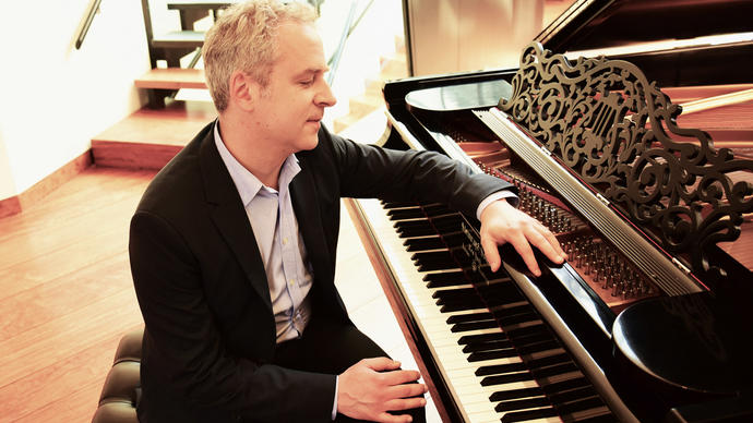 Jeremy Denk sits at a piano with one arm resting on it