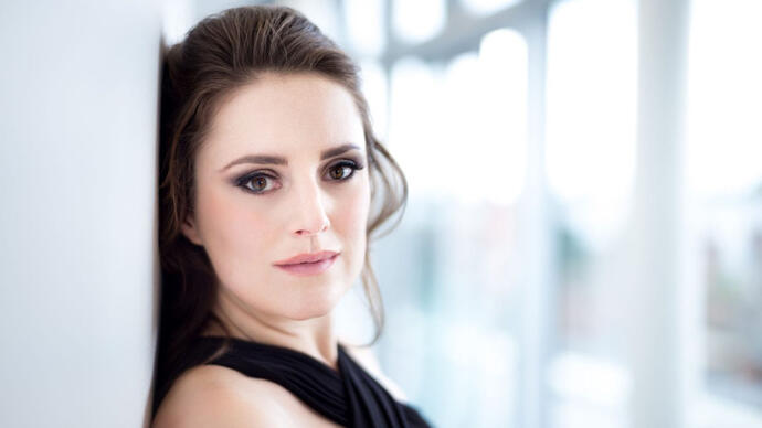 Soprano Louise Alder looks at the camera, leaning against a white wall