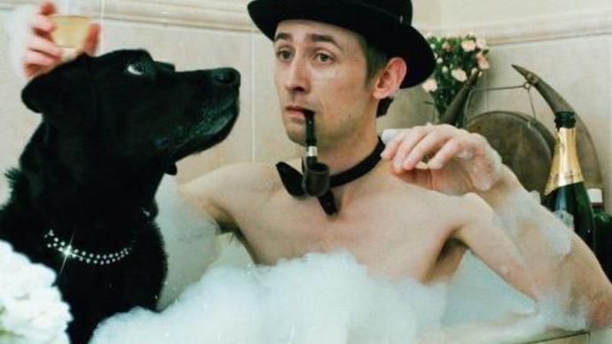 Photo of a man wearing a black hat sitting in a bath full of bubbles with a black dog