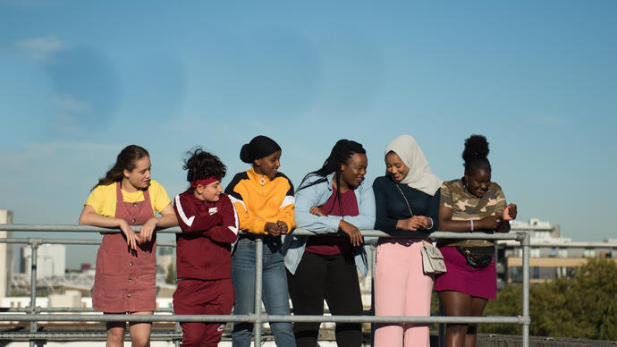 A group of six young girls stand in a row on what looks like the roof of a building. They are leaning on some railings and there's a clear blue sky above them.