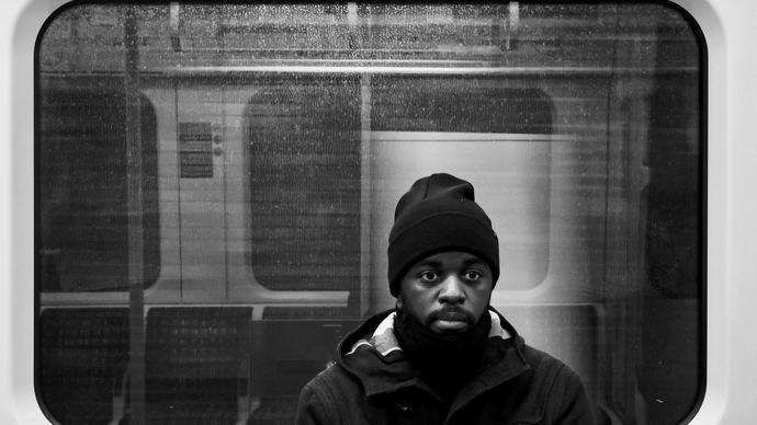 black and white photo of Alpha Mist sitting in a tube carriage, he is wearing a big coat and a beanie hat, with a neutral expression