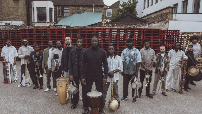 Balimaya Project standing in a triange formation with their instruments at their feet, in front of stacked red crates.