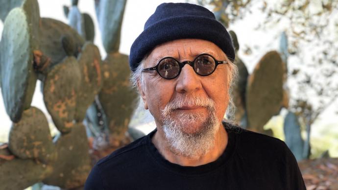 Charles Lloyd in a black beanie hat and small round sunglasses, standing in front of a large cactus