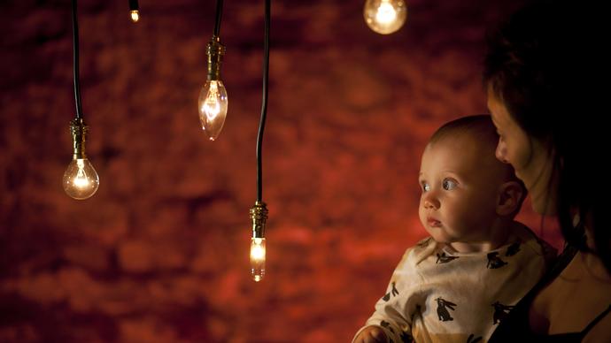 A baby in the arms of their mother look at hanging tungsten style light bulbs, bathed in a warm glow. Behind them is a red exposed brick wall. The mother is looking intently at the child.