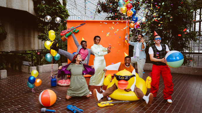 A party of people in front of an orange screen wearing party hats, dancing with confetti and inflatables.