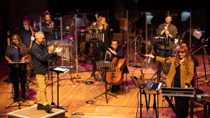 Hannah Peel performing with Paraorchestra. From behind a keyboard on the left of the stage she is gesturing to the ensemble who are applauding.